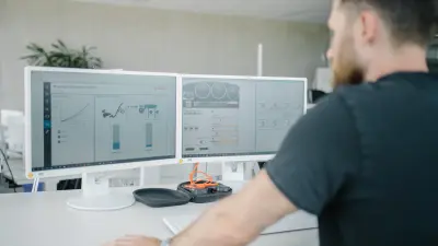 Zwei Computer-Bildschirme stehen nebeneinander auf einem Schreibtisch in einer Büroumgebung. Darauf sind Messungen und Diagramme zu erkennen. Dominic ist unscharf im Vordergrund zu sehen, sein Blick ist auf die Bildschirme gerichtet.