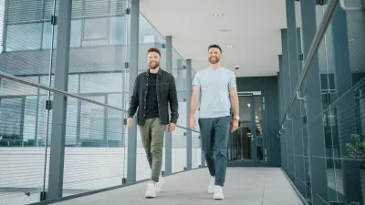 Dominic (links) und Andreas (rechts) laufen lächelnd einen Gang entlang. Der Gang ist auf beiden Seiten verglast, im Hintergrund befindet sich eine Tür, die in ein Gebäude führt.