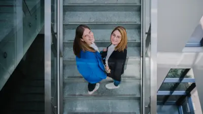 : Angela und Nicole stehen in einem Treppenhaus in einem Bürogebäude und blicken nach oben. Nicole steht links von Angela.