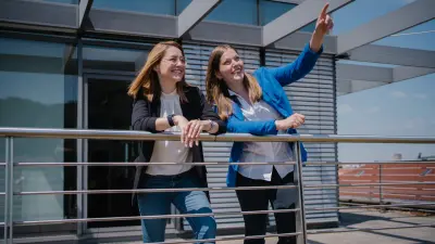 Angela und Nicole befinden sich im Außenbereich auf einer Terrasse. Sie lehnen beide an einem Geländer. Beide blicken nach vorne und lächeln. Nicole steht rechts von Angela und zeigt mit ihrer linken Hand in die Ferne.