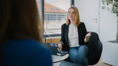 Angela sitzt in einem schwarzen Sessel an einem runden kleinen Tisch, auf dem ein Laptop steht. Sie hebt ihre linke Hand an und lächelt in Richtung Nicole, die ihr gegenübersitzt.