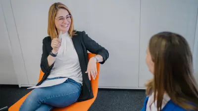 Angela sitzt auf einem orangenen Stuhl und lächelt in Nicoles Richtung, die neben ihr sitzt. Sie hält einen Kugelschreiber in ihrer rechten Hand und zeigt damit nach oben.