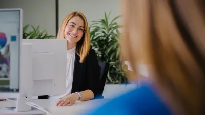 Angela sitzt an einem Schreibtisch in einer Büroumgebung. Sie blickt an ihrem Bildschirm vorbei und lächelt Nicole im Vordergrund an, die ihr gegenüber sitzt.