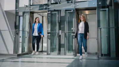 Nicole und Angela verlassen gleichzeitig zwei verglaste Fahrstühle, die sich nebeneinander in einem Treppenhaus befinden. Nicole blickt lächelnd zu Angela, Angela lächelt in die Kamera. Beide tragen einen Laptop unter dem Arm.