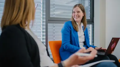 Nicole sitzt auf einem orangen Stuhl mit ihrem Laptop auf dem Schoß. Sie lächelt in Angelas Richtung, die neben ihr sitzt.