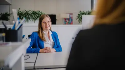 Nicole sitzt mit verschränkten Armen an einem Schreibtisch in einer Büroumgebung. Sie lächelt Angela im Vordergrund an, die ihr gegenübersitzt.
