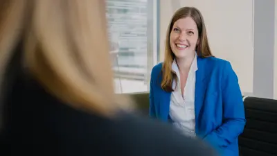 Nicole befindet sich in einer Büroumgebung und sitzt auf einem schwarzen Sessel. Sie trägt einen blauen Blazer und lächelt in Richtung von Angela, die ihr gegenübersitzt.