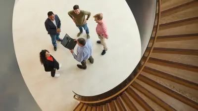 Vier Männer und eine Frau stehen vor einer Wendeltreppe und lachen. Der Blick geht von oben auf die Gruppe herab. Ein Mann hält einen Laptop in der Hand.