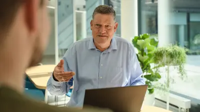 Ein Mann sitzt hinter seinem Laptop. Er gestikuliert mit den Händen und erklärt etwas. Im Vordergrund ist sein Zuhörer unscharf zu sehen.