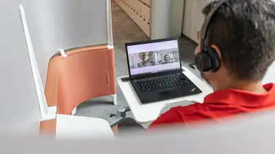 Mustafa lächelt in die Kamera seines Laptops, während er mit einer Kollegin im Online-Meeting ist. Er sitzt in einer Ruheecke.