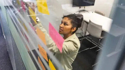 Sarah klebt beschriebene Haftnotizzettel an eine Glasscheibe in einem Meetingraum.