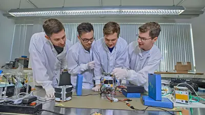 Vier Studenten stehen vor einem Tisch, auf dem ein kleiner Satellit liegt.