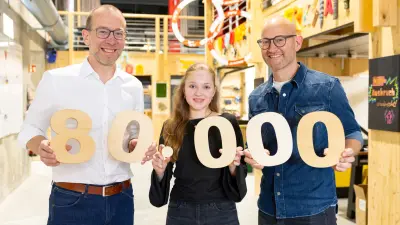 Zwei Männer und ein junges Mädchen stehen nebeneinander und halten große Holzziffern in die Kamera, die die Zahl 80.000 ergeben. Alle drei lächeln, der Hintergrund zeigt eine moderne Holzkonstruktion.