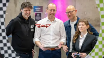Vier Personen – drei Männer und eine junge Frau – stehen zusammen. In der Mitte schwebt eine kleine, rote Drohne, die von einem der Männer mit einem Tablet gesteuert wird, während die junge Frau eine Fernbedienung hält. Der Hintergrund ist mit einem Schachbrettmuster und pinker Beleuchtung gestaltet.