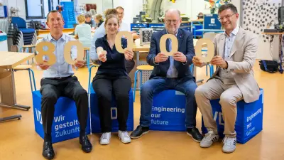 Vier Personen, drei Männer und eine Frau, sitzen nebeneinander auf blauen Würfeln mit der Aufschrift "Wir gestalten Zukunft" und "Engineering our future". Sie halten große hölzerne Ziffern in die Kamera, die die Zahl 80 000 ergeben, und lächeln. Die Szene findet in einem hellen Klassenraum oder einer Werkstatt statt.