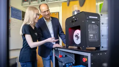 Ein Mann und eine junge Frau stehen lächelnd neben einem schwarzen 3D-Drucker. Die Frau zeigt auf einen Teil der Maschine, während sie dem Mann etwas erklärt.