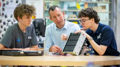 Ein Mann in einem hellblauen Hemd sitzt an einem Holztisch zwischen zwei jüngeren Männern und erklärt ihnen ein technisches Gerät, das wie ein Solarmodul aussieht. Alle drei Personen sind konzentriert auf das Gerät gerichtet.