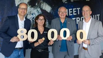 Vier Personen – drei Männer und eine Frau – halten große Holzziffern, die die Zahl 80.000 bilden, und lächeln in die Kamera. Im Hintergrund ist ein Banner mit einem Astronauten im Weltall und den Logos von DLR, Universität Stuttgart und IRS zu sehen.