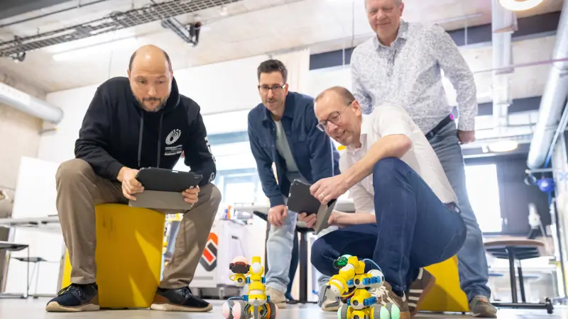 Vier Männer beobachten in einem hellen, modernen Raum zwei kleine, selbstgebaute Roboter auf dem Boden.